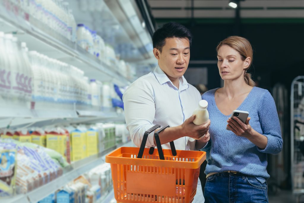Asian couple using smartphone in supermarket with shopping cart choosing products while grocery store